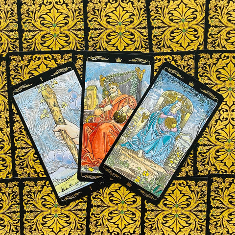 Tarot kortos su juodai auksinėmis ornamentuotomis nugarėlėmis: trys atvertos kortos (Lazdos, Karalius, Karalienė Pentaklių) elegantiškai išskleistos virš raštuoto fono