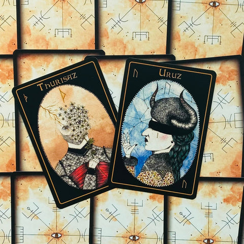 Tarot kortos juodais rėmais su dviem centrinėmis iliustracijomis (Uruz, Thurisaz), atviros virš foninių kortų