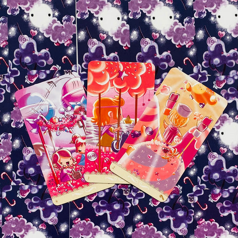 Tarot kortos trijų kortų išklotinė su ryškiomis rožinėmis saldumynų tematikos iliustracijomis ant tamsiai mėlyno fono