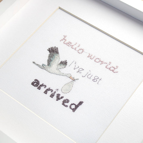 Siuvinėtas paveikslas baltame rėme su skraidančiu gandru, maišeliu ir ranka siūtu užrašu 'hello world'