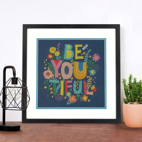Siuvinėtas paveikslas rėme tamsiai mėlynas su spalvotomis „BE YOU TIFUL“ raidėmis ir gėlių motyvais, stilizuotas ant stalo prie lempos ir sukulentų