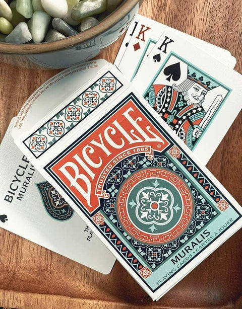 Bicycle kortų kaladė Muralis su baltai-oranginėmis ir žalsvomis detalėmis, atversta ant medinio stalo su matomais karaliais