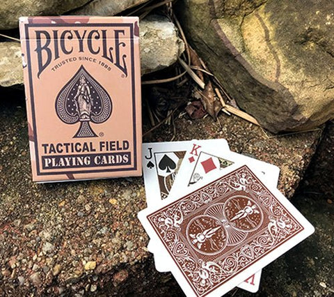Bicycle Tactical Field kortos rudos dėžutės ir atvertos kortos su ornamentine nugarėle padėtos ant akmenų lauke