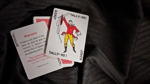 Tally‑Ho Joker kortos, dvi persidengiančios baltos kortos su raudonai‑geltonu jokeriu ir tekstu ant juodo audinio fono