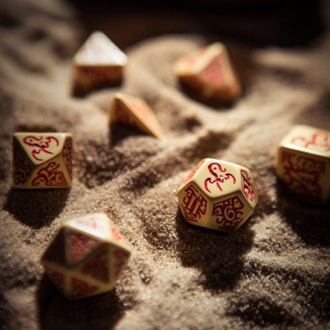 RPG kauliukai kreminiai su raudonais ornamentais, artimas D20 fokusas ant smėlingo fono pabrėžia detalias žymes ir tekstūrą