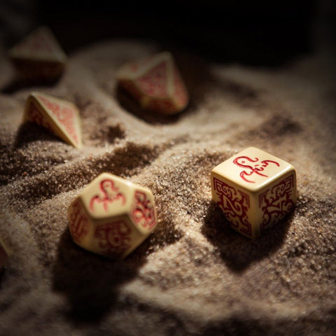 RPG kauliukai kreminės spalvos su raudonais ornamentais, arti rodomas kubas ir daugiasienis kauliukas ant smėlio fono
