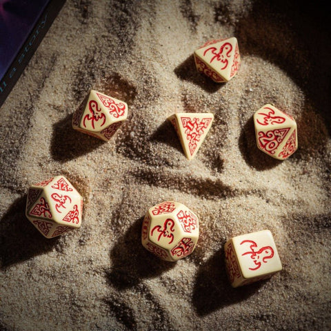 RPG kauliukai kreminės spalvos su raudonomis ornamentinėmis graviruotėmis, 7 dalių komplektas ant smėlio, artimas kampas