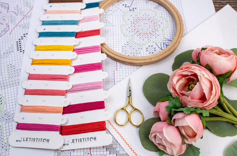 Siuvinėjimo siūlai LETISTITCH ryškių rožinių, raudonų ir geltonų atspalvių kortelėje, šalia medinio lankelio, auksinių žirklių ir rožių dekoracijos