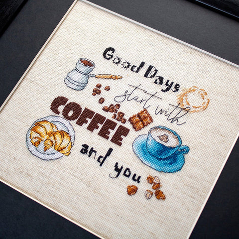 Siuvinėtas kryželiu paveikslas Coffee and you su mėlynu kavos puodeliu, džezva, kruasanu ir kavos pupelėmis, artimas kampinis kadras su rėmu