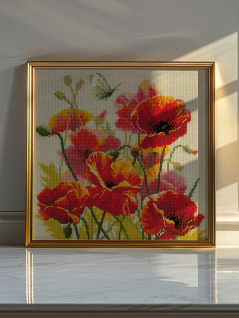 Siuvinėjimas rėmelyje su ryškiomis raudonomis aguonomis ir žalsvu drugeliu, 40×40 cm, padėtas ant stalo saulės šviesoje