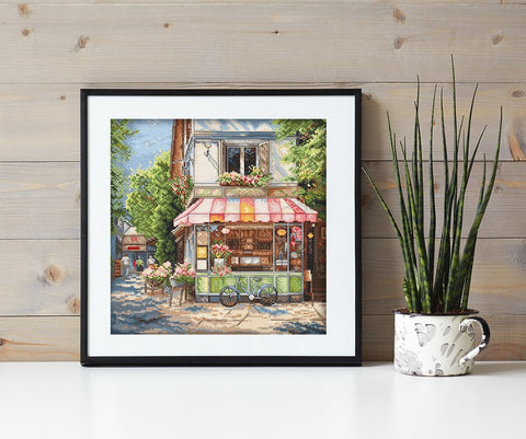 Kryželių siuvinys kvadratinis kavinės motyvas, juodas rėmas 30×30 cm, stilingai eksponuotas ant medinės sienos