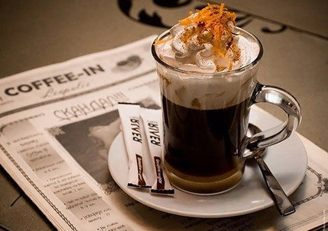 Kava espresso taurėje su plakta grietinėle ir tarkuota apelsino žieve, ant baltos lėkštutės su cukraus pakeliais ir šaukšteliu