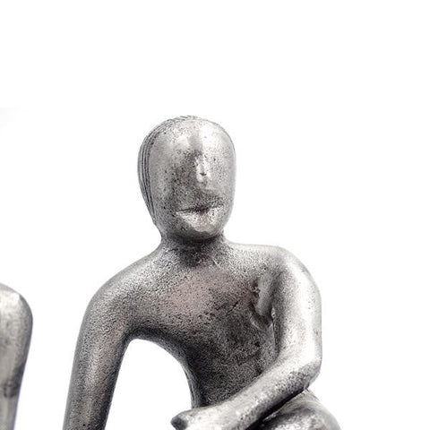 Skulptūra – sidabro spalvos sėdinti dekoratyvinė figūra, arti apkirpta, matomas viršutinis kūnas ir ranka iš dešinės kampo