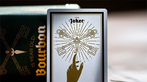 Kortų kaladė su balta Joker korta ir auksiniu raktų dizainu, arti, priešais juodą Bourbon dėklą.