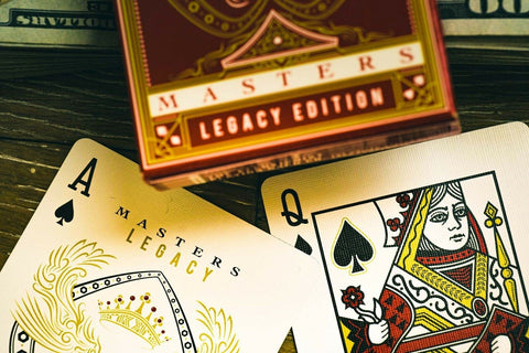Kortos Masters Legacy Edition kreminės su auksiniais akcentais, artimas Ace ir Queen kortų kampų kadras su tūrinio dėklo pakeliu viršuje