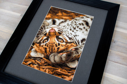 Siuvinėtas leopardas rudo-baltos spalvos, kampu įrėmintas juodu mediniu rėmu su pilku paspartinimu, detali priartinta perspektyva