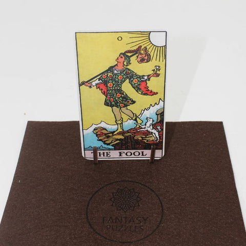 Tarot korta The Fool, Rider–Waite stiliaus, stačia ant mažo medinio stovo, eksponuota ant rudo Fantasy Puzzles kilimėlio, matomas priekis