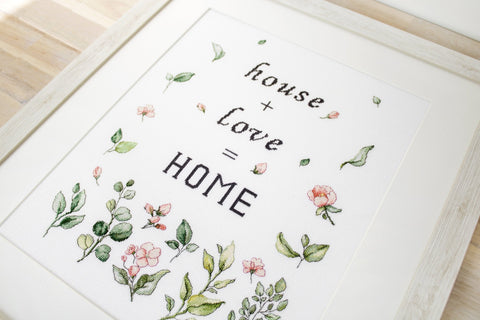 Siuvinėtas paveikslas su užrašu "house + love = HOME" ir švelniomis rožinėmis gėlėmis, baltame mediniame rėme, fotografuotas iš kampo