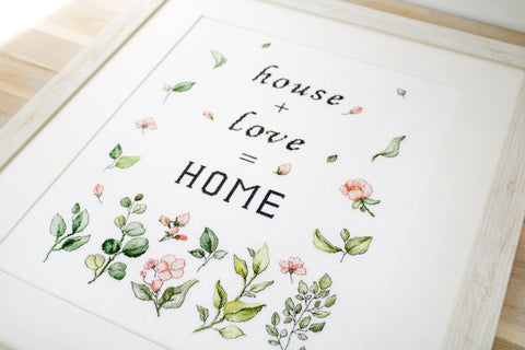Siuvinėtas rėminis paveikslas su juodu užrašu HOME ir smulkiais rožiniais bei žalių lapų motyvais, šviesaus medžio rėme, nufotografuotas kampu