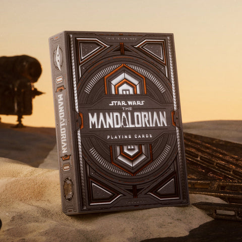 Mandalorian žaidimo kortų kaladė, sidabrinės ir varinės spalvų metalizuota pakuotė priekinio kampo perspektyvoje ant smėlio, akcentuojant įmantrų dekorą
