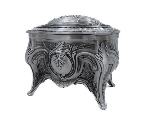 Papuošalų dėžutė sidabro spalvos, antikvarinė metalinė su reljefiniais ornamentais, matoma iš priekinio kampo ir pabrėžianti tris kojas