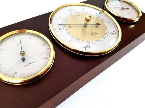 meteorologinė stotelė ant tamsiai rudos medienos su trimis auksuotais ciferblatais — barometras, termometras, drėgmėmatis, kampinis artimas planas