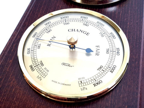 Barometras Fischer su aukso rėmu ir baltu ciferblatu ant tamsaus medinio pagrindo, rodyklė rodo Change
