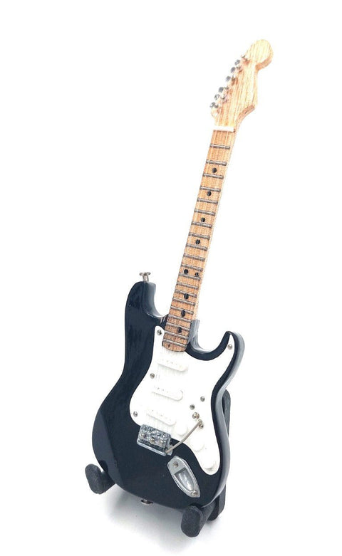 Elektrinė gitara juoda su balta apsaugos plokštele, kleviniu grifu ir trijų pikapų konfiguracija, trijų ketvirčių priekio kampu ant stovelio