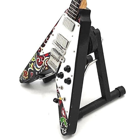 Elektrinė gitara Flying V su balta priekyje ir spalvotu gėlių raštu, chromuotais pikapais, kampinis vaizdas ant juodo stovo