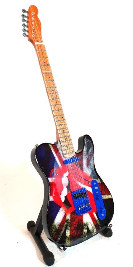 Elektrinė gitara su Union Jack korpusu, kleviniu kaklu ir mėlynais paėmėjais, stovinti ant stovelio pasviru kampu