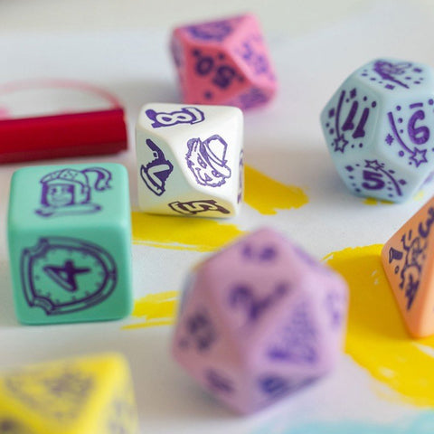 Kauliukai RPG rinkinyje pasteliniai (mėtiniai, rožiniai, violetiniai), daugiašonės su smulkiais iliustruotais simboliais, arti ant geltono potėpio