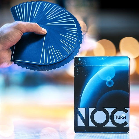 Kortos NOC Turn mėlynos plonos, rankoje platus vėduoklės fanas ir stilinga dėžutė ant stalo, pabrėžiant jų tekstūrą ir lankstumą