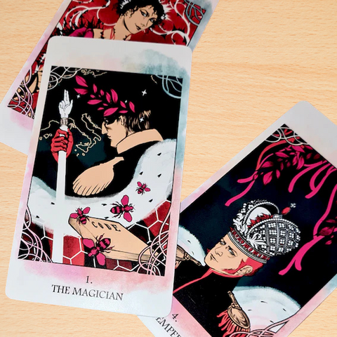 Tarot kortos su The Magician dizainu, pastelinėmis rožinėmis detalėmis, kampu užfiksuotos ant medinio fono, matomas ir Imperatorius