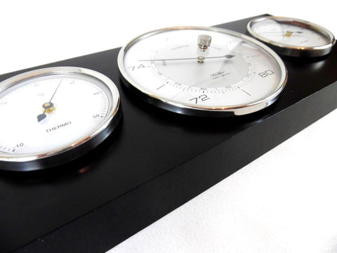 Oras stotelė su juodu mediniu pagrindu ir trimis chromuotais baltais ciferblatais, kampinis priartinimas pabrėžia barometro centrą ir termometro skalę