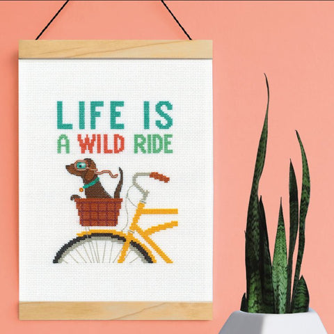 Kryželiu siuvinėtas paveikslas „Life Is a Wild Ride“ su geltonu dviračiu ir šuniuku krepšyje, kabantis ant ryškios rožinės sienos