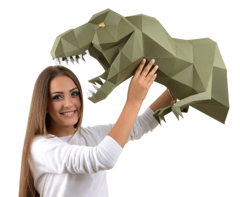 Popierinė T‑rex galva žalsva, didelė geometrinė sieninė dekoracija, modelis laiko ją parodydamas tikrą dydį ir formą