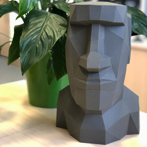 Busto skulptūra, pilka geometrinė moderni, apie 30 cm aukščio, nufotografuota iš priekio ant stalo šalia žalsvo vazonėlio