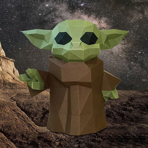 Baby Yoda figūrėlė iš kartono su žalia poligonine galva ir rudu chalatu, stovi ant uolėtos kosminės scenos