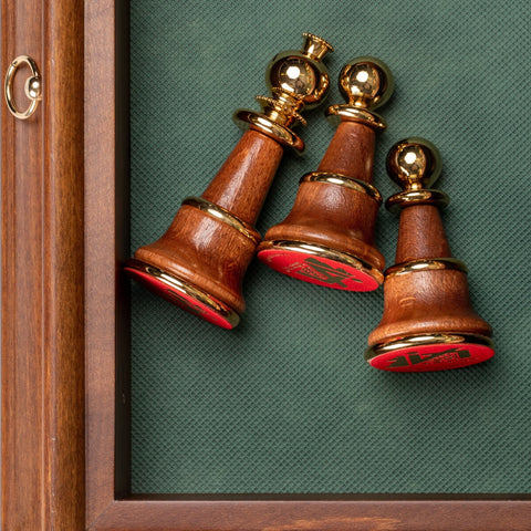 Šachmatų figūros iš tamsaus medžio su auksinėmis detalėmis ir raudonomis vilnonėmis apačiomis, išdėstytos šalia medinio rėmo ant žalsvo audinio