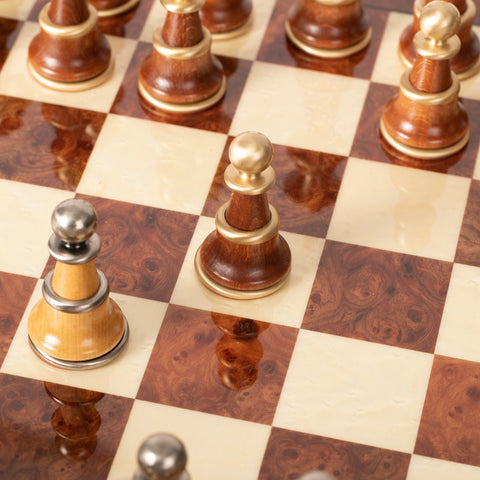 Šachmatų rinkinys – artimas kampinis kadras su riešuto ir kreminės spalvų lenta, medinės figūrėlės su aukso ir sidabro žiedais, akcentuojant blizgantį apdailos paviršių