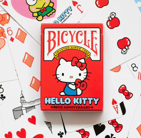 Bicycle žaidimų kortos Hello Kitty 50‑mečio leidimas, ryškus raudonas dėklas centre, kortos išdėstytos fone kaip galerijos variantas