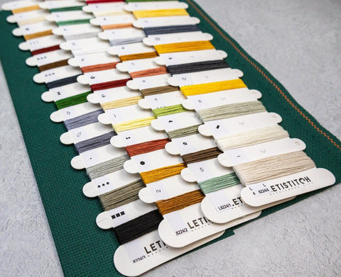 Siuvinėjimo siūlai ant tamsiai žalsvos Aida drobės, išdėstyti įstrižai kortelėse su žemės, geltonų ir neutralių atspalvių palete