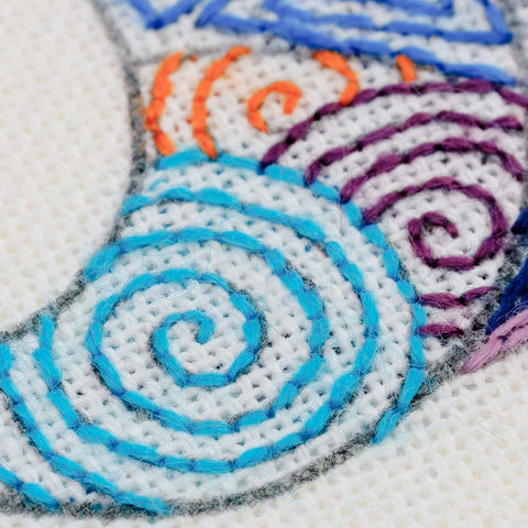 Siuvinėjimo rinkinys, artimas planas: mėlynos, violetinės ir oranžinės spiralės ant baltos Aida drobės, matomas siūlių reljefas