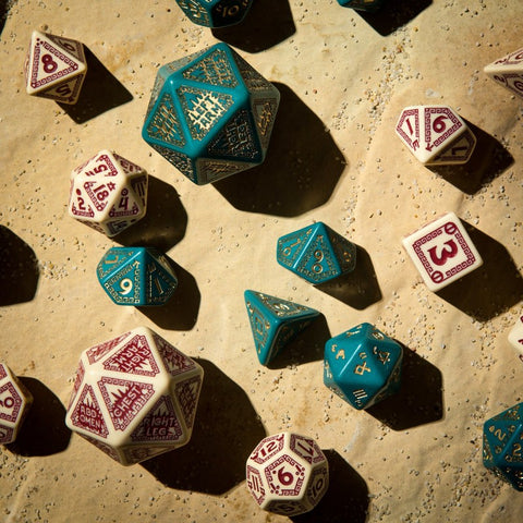 RPG kauliukai: žalsvai mėlynas metalinis rinkinys su auksinėmis graviruotėmis šalia kreminio rinkinio su burgundiškomis įrašomis