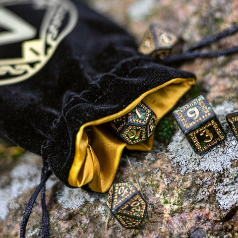 RPG kauliukai iš žalvario su žaliais akcentais, išlindę iš juodo aksominio maišelio su auksiniu pamušalu