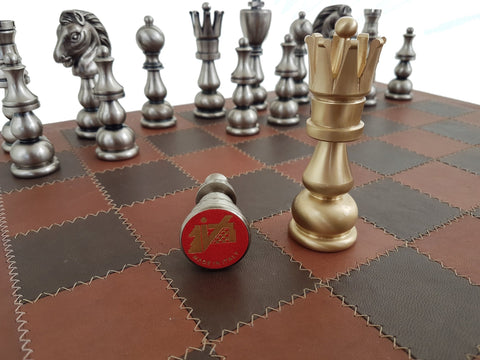 Šachmatai su auksine karaliene stovinčia prie nukritusio sidabrinio karaliaus ant siūtuotos rudos odinės lentos, matomas Made in Italy ženklas