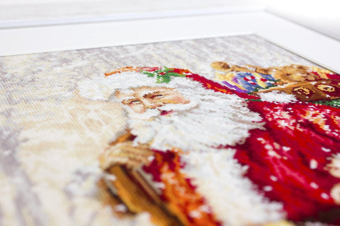 Kryželiu siuvinėtas Kalėdų Senelis rėminis paveikslas iš arti, paryškintos siūlų tekstūros, ryškios raudonos ir baltos spalvos