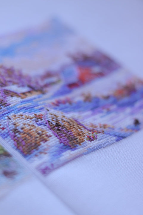 Siuvinėjimas kryželiu, artimas planas: pastelinės violetinės ir mėlynos siūlės ant baltos drobės, išryškėjusi rašto tekstūra