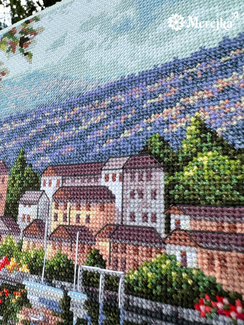 Kryželiu siuvinys Merejka, artimas levandų laukų ir upės pakrantės namų fragmentas su ryškia violetine ir žalia siūlų tekstūra