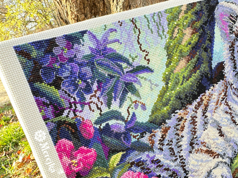 Kryželių siuvinėjimo rinkinys Marijke, arti kampo fokusas su violetinėmis ir rožinėmis gėlėmis bei dryžuotu kačiuko kailiu, fotografuotas lauke saulės apšvietime